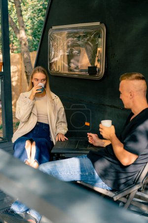 Foto de Una pareja joven un hombre y una chica toman té y charlan en la terraza cerca de una autocaravana en una caravana - Imagen libre de derechos