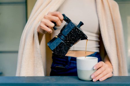Téléchargez les photos : Gros plan de la main d'une femme tenant un verre et versant du café fraîchement infusé d'une cafetière sur une terrasse avec des fleurs - en image libre de droit
