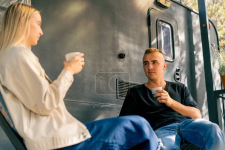 Foto de Un chico guapo joven y una chica están sentados en la terraza de una autocaravana en una caravana bebiendo té y hablando con entusiasmo - Imagen libre de derechos