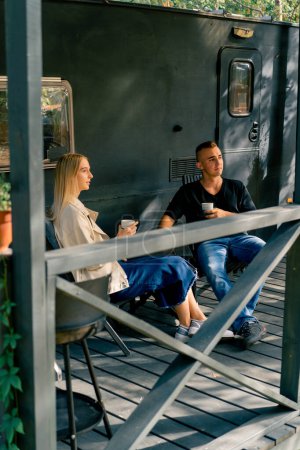 Téléchargez les photos : Un jeune beau mec et une fille sont assis sur la véranda d'un camping-car dans un camping-car boire du thé et parler avec enthousiasme - en image libre de droit