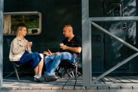 Téléchargez les photos : Un jeune beau mec et une fille sont assis sur la véranda d'un camping-car dans un camping-car boire du thé et parler avec enthousiasme - en image libre de droit