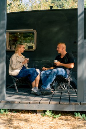 Photo for A young handsome guy and a girl are sitting on the veranda of a motorhome in a camper drinking tea and talking enthusiastically - Royalty Free Image