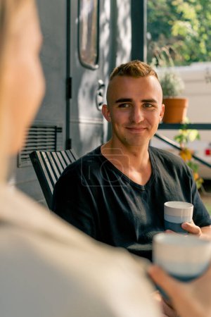 Téléchargez les photos : Gros plan d'un jeune homme assis sur la terrasse près d'un mobil-home buvant du thé et écoutant attentivement sa petite amie - en image libre de droit