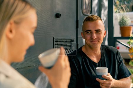 Photo for Close-up shot of a young guy sitting on the terrace near a mobile home drinking tea and listening carefully to his girlfriend - Royalty Free Image