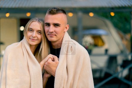 Téléchargez les photos : Un couple mignon amoureux une fille et un mec sont enveloppés dans une couverture et câlins dans le contexte d'une maison sur roues en glamping - en image libre de droit