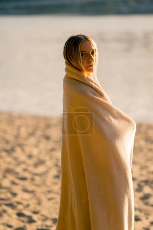 Téléchargez les photos : Une fille solitaire enveloppée dans une couverture douce regarde le coucher de soleil sur la rive sablonneuse de la rivière et sourit - en image libre de droit