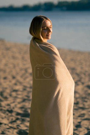 Photo for A lonely girl wrapped in a soft blanket watches the sunset on the sandy bank of the river and smiles - Royalty Free Image