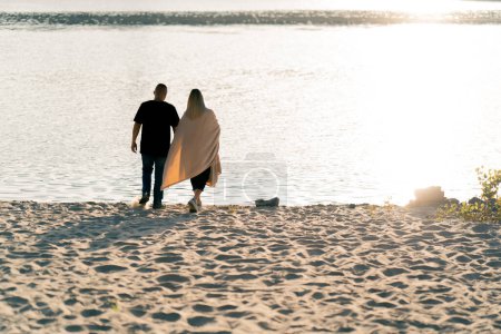 Téléchargez les photos : Couple amoureux tenant les mains couvertes d'une couverture et marchant le long de la rive sablonneuse vers la rivière au coucher du soleil - en image libre de droit