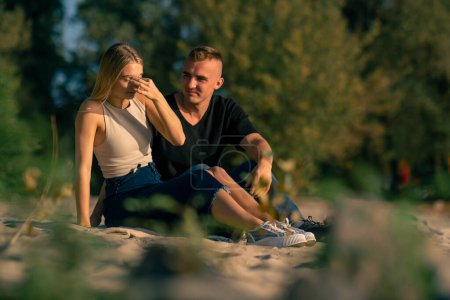 Foto de Un joven pelea con su novia que se aleja de él ofendido mientras está sentado en la orilla arenosa del río - Imagen libre de derechos