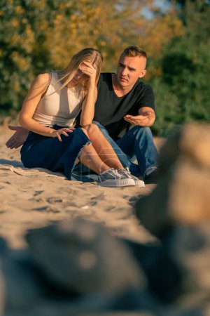Téléchargez les photos : Un jeune homme se dispute avec sa petite amie qui se détourne de lui offensé alors qu'il est assis sur le rivage sablonneux de la rivière - en image libre de droit
