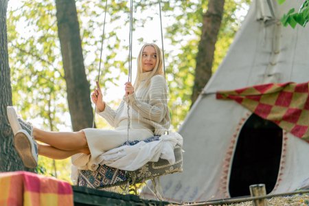 Photo for A young girl in beautiful clothes smiles and rides on a swing in the forest against the background of a tree house - Royalty Free Image