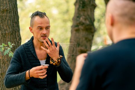 Téléchargez les photos : Maître de thé masculin extravagant avec des bracelets traditionnels à la main racontant aux invités le thé lors d'une cérémonie dans le jardin - en image libre de droit