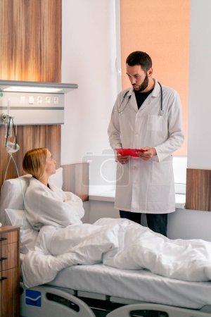 Foto de Durante una mañana, el médico de cabecera visita a su paciente en la sala a un monitor de la dinámica de su tratamiento. - Imagen libre de derechos