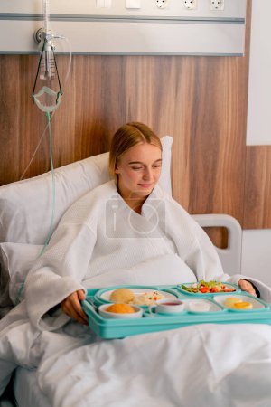 Téléchargez les photos : Une fille allongée dans une chambre d'hôpital est amenée plateau avec un délicieux aliment pour renforcer sa force et se remettre de la maladie - en image libre de droit