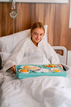 Photo for A girl lying in a hospital room is brought tray with a delicious food to strengthen her strength and recover from illness - Royalty Free Image