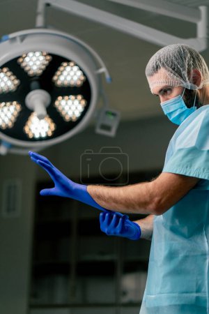 Photo for A doctor in the operating room in a protective uniform and a special headdress puts on gloves before an operation in an operating room - Royalty Free Image