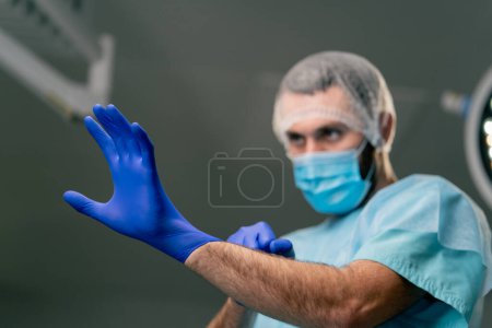 Photo for Close-up shot of a surgeon in uniform and medical mask putting on gloves in operating room while preparing for a surgery - Royalty Free Image