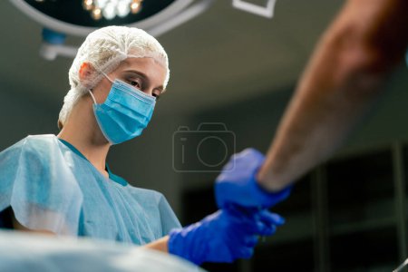 Photo for A girl assistant surgeon in an uniform and a medical mask passes scalpel to her colleague during an operation - Royalty Free Image