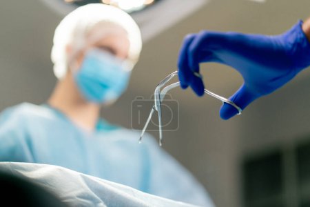 Photo for A girl assistant surgeon in an uniform and a medical mask passes scalpel to her colleague during an operation - Royalty Free Image