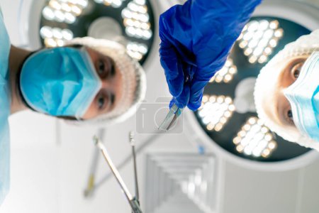 Foto de Cirujanos profesionales en uniforme sobre el fondo de lámparas dobladas sobre un paciente acostado en la mesa de operaciones para realizar una operación - Imagen libre de derechos