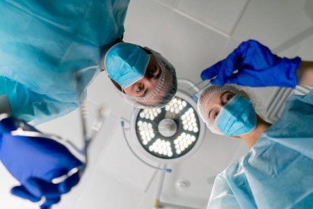 Téléchargez les photos : Chirurgiens professionnels en uniforme sur fond de lampes pliées sur un patient allongé sur la table d'opération pour effectuer une opération - en image libre de droit