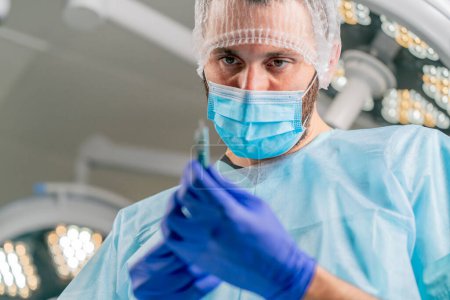 Photo for Close-up shot of a surgeon in a protective headdress and a medical mask holding scalpel in his hands and looking at it carefully - Royalty Free Image