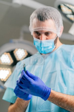 Foto de Primer plano de un cirujano con un tocado protector y una máscara médica sosteniendo el bisturí en sus manos y mirándolo cuidadosamente - Imagen libre de derechos