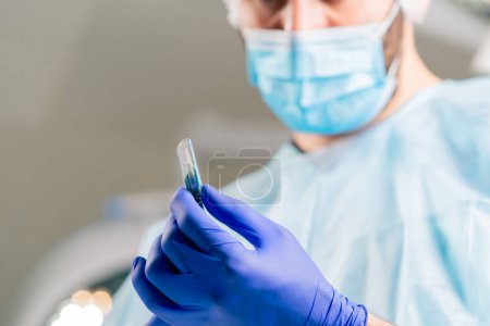 Photo for Close-up shot of a surgeon in a protective headdress and a medical mask holding scalpel in a his hands and looking at it carefully - Royalty Free Image