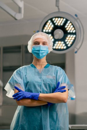 Foto de Retrato de una anestesióloga con uniforme y máscara médica en quirófano con los brazos cruzados delante de ella - Imagen libre de derechos