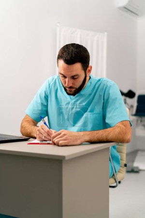 Foto de Un médico con uniforme médico se sienta en un consultorio médico en su escritorio con una computadora portátil y llena los papeles. - Imagen libre de derechos