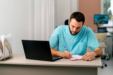 Téléchargez les photos : Un médecin en uniforme médical est assis dans un bureau médical à son bureau avec un ordinateur portable et remplit des papiers - en image libre de droit