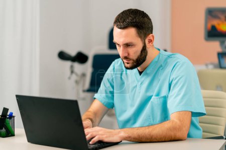 Téléchargez les photos : Un médecin barbu consulte en ligne un patient à l'aide d'un ordinateur portable dans son cabinet médical - en image libre de droit