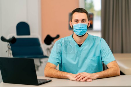 Foto de Retrato de un médico con uniforme y una máscara médica sentado en su escritorio en su oficina en una clínica - Imagen libre de derechos