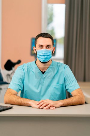 Photo for Portrait of a male doctor in a uniform and a medical mask sitting at his desk in his office in a clinic - Royalty Free Image