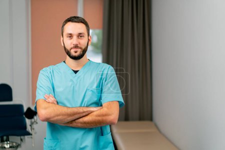 Photo for Portrait of a male doctor with a beard stands in medical office with his hands folded in a front of him and looks straight - Royalty Free Image
