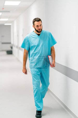 Téléchargez les photos : Un grand médecin avec une barbe marche le long du couloir de l'hôpital avec un dossier de documents - en image libre de droit
