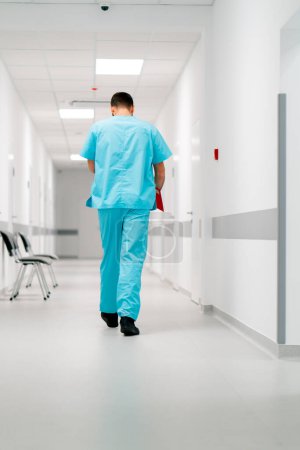 Photo for A tall male doctor with a beard walks along the hospital corridor with a folder of documents - Royalty Free Image