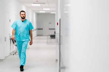 Téléchargez les photos : Un grand médecin avec une barbe marche le long du couloir de l'hôpital avec un dossier de documents et entre dans son bureau - en image libre de droit