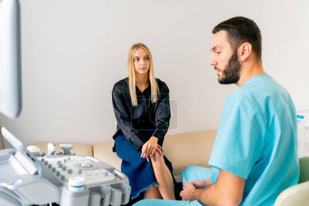 Photo for A girl patient sits on a gurney in the ultrasound diagnostic room after the procedure and listens carefully to a doctor - Royalty Free Image