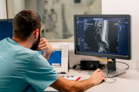 Foto de Un radiólogo se sienta en una mesa detrás de un monitor de computadora y examina una imagen de resonancia magnética durante un examen de un paciente. - Imagen libre de derechos
