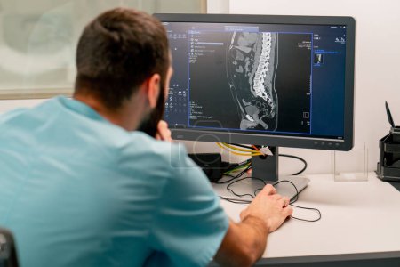 Photo for A radiologist sits at a table behind a computer monitor and examines a magnetic resonance imaging image during an examination of a patient - Royalty Free Image