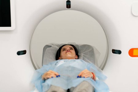 Photo for A girl in a medical gown lies on a movable couch of a magnetic resonance imaging machine and prepares for a diagnosis - Royalty Free Image