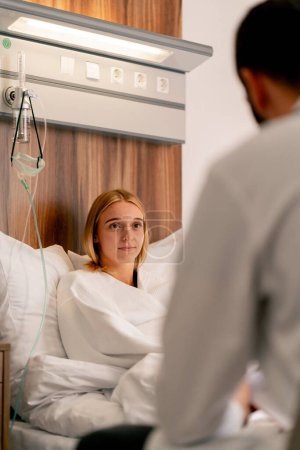 Foto de Una niña enferma en bata de baño se encuentra en la sala médica de un hospital hospitalario y describe su estado de salud a su médico que atiende - Imagen libre de derechos