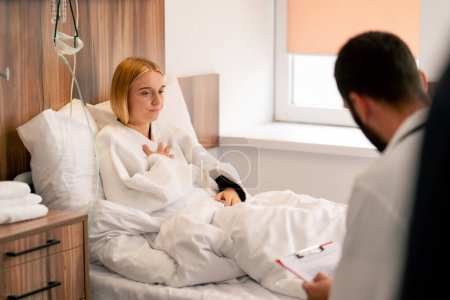 Téléchargez les photos : Un médecin vêtu d'un manteau blanc s'assoit près du lit d'une fille malade dans un service et écoute ses plaintes concernant sa santé. - en image libre de droit