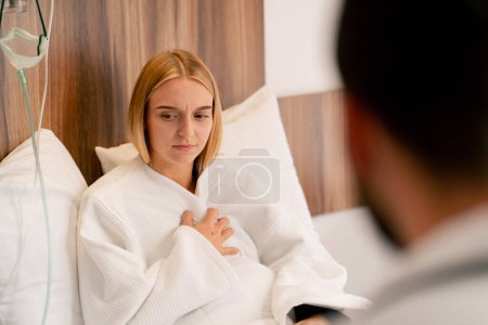 Téléchargez les photos : Une fille malade est allongée dans une chambre d'hôpital et tient sa poitrine avec sa main et tousse près du médecin - en image libre de droit