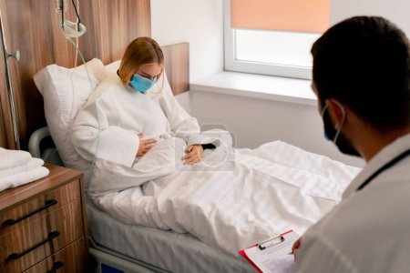 Foto de Una niña en una habitación de hospital con una máscara médica sostiene su estómago con la mano en un dolor y escucha al médico con sus recomendaciones - Imagen libre de derechos