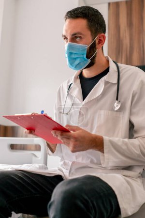 Téléchargez les photos : Gros plan d'un médecin portant un masque médical et une blouse d'hôpital notant les plaintes du patient dans les antécédents médicaux - en image libre de droit