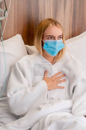 Téléchargez les photos : Gros plan d'une fille malade portant un masque médical couchée dans un service toussant et se couvrant la bouche avec sa main - en image libre de droit