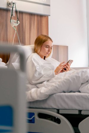 Photo for A sad girl in a bathrobe lies in a hospital room and sadly looks at her phone while waiting for her doctor - Royalty Free Image