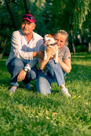 Téléchargez les photos : Portrait d'une famille heureuse un homme adulte et une femme en promenade avec un chien de la race Jack Russell terrier dans le parc de la ville étreignant animal - en image libre de droit
